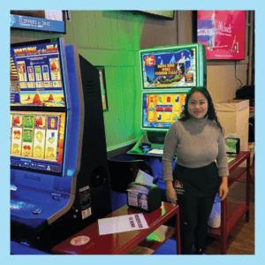 Inside of Leo's Lair, we see a smiling Asian woman with long wavy black hair wearing a gray turtleneck and an Oregon Lottery apron over black trousers. She is standing between Oregon Lottery video lottery terminals inside of a bar, decorated with wood paneling.