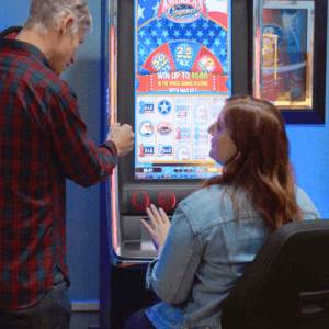 Red-haired woman in a denim jacket sitting in front of a Video Lottery game machine. Standing next to her is a man with short gray hair in a plaid shirt. They are talking and smiling.