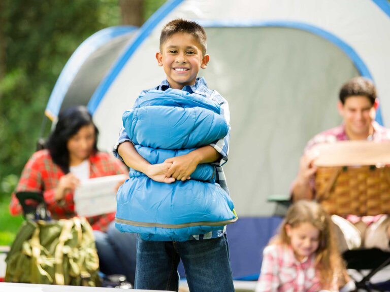 A happy family in front of a tent; the son holds a rolled sleeping bag in the foreground.