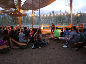 Campers sit on outdoor benches and listen to a man who sings while playing guitar.