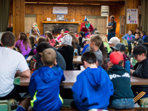 A large group of campers assemble at tables indoors for a group activity.